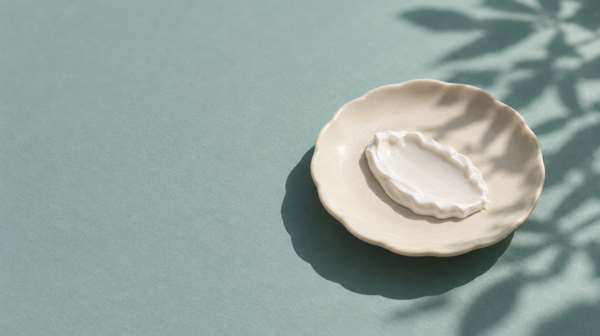 A dollop of thick white mineral sunscreen on a cream ceramic dish, with the shadow of a leaf cast across it in bright daylight.