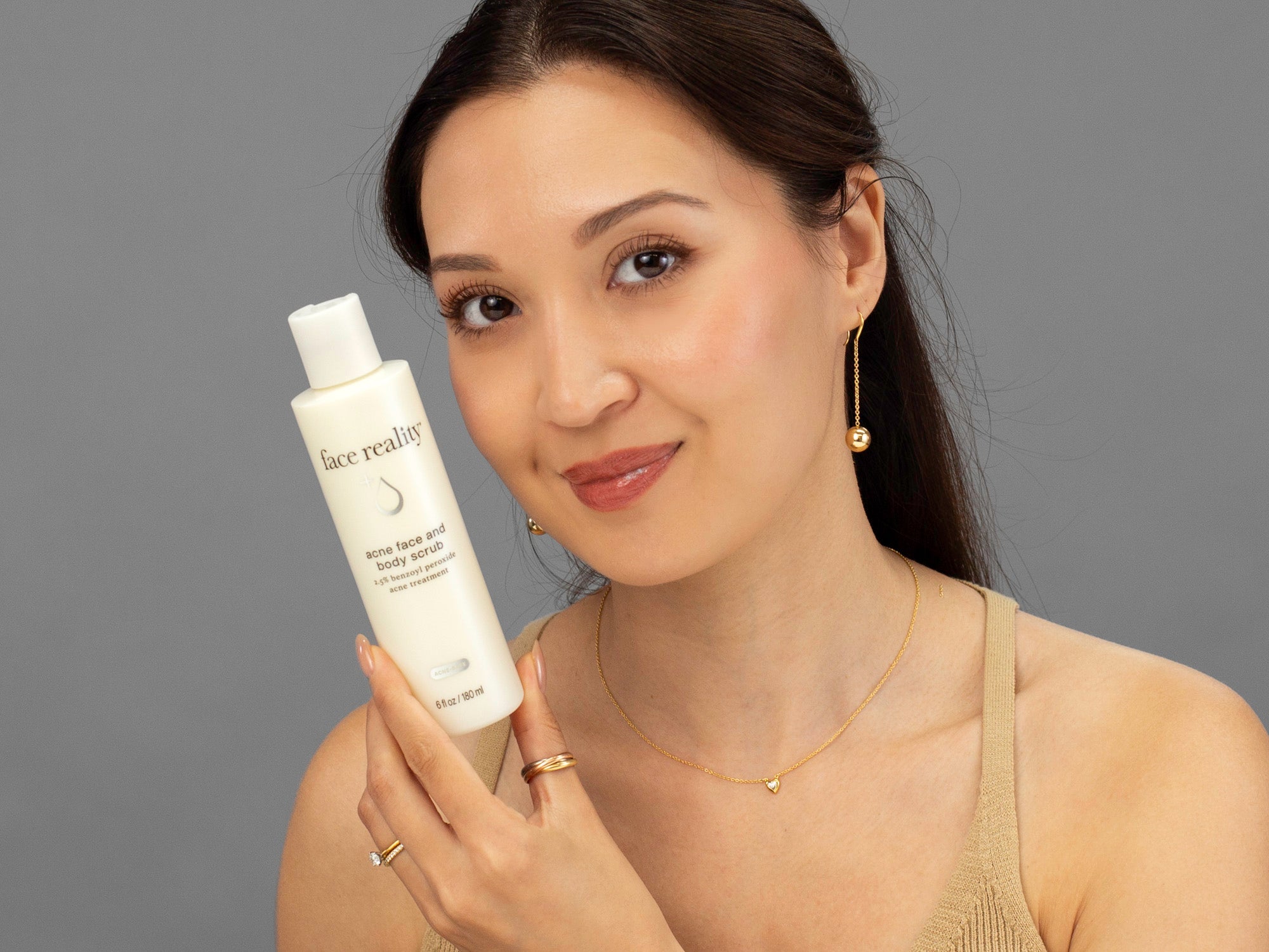 Laziza from LAZ Skincare holding a bottle of Face Reality Acne Face & Body Scrub up to her face