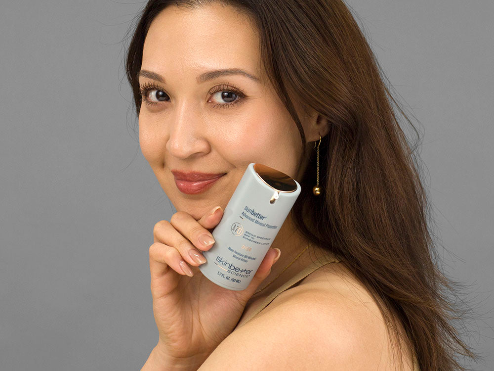 Laziza from LAZ Skincare holding a bottle of SkinBetter Science sunbetter SHEER SPF 70 Sunscreen Lotion up to her face