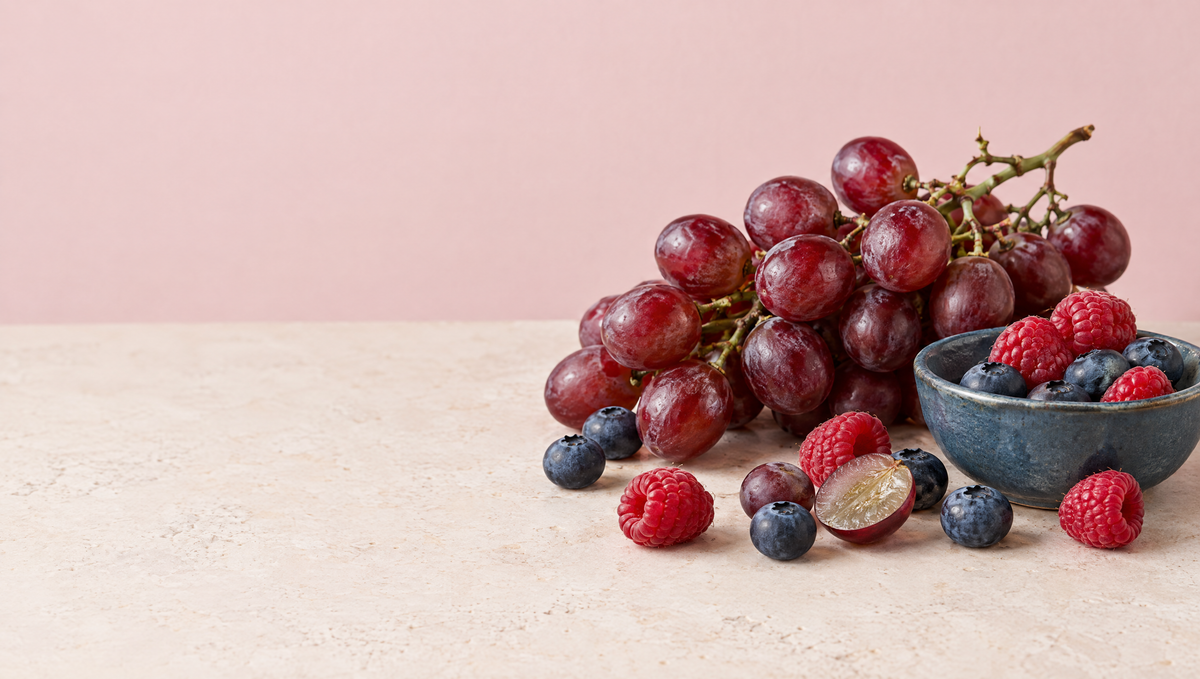 Red grapes on the vine with blueberries, raspberries and a halved grape on a warm stone surface against a blush backdrop.