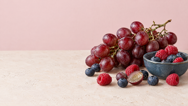 Red grapes on the vine with blueberries, raspberries and a halved grape on a warm stone surface against a blush backdrop.