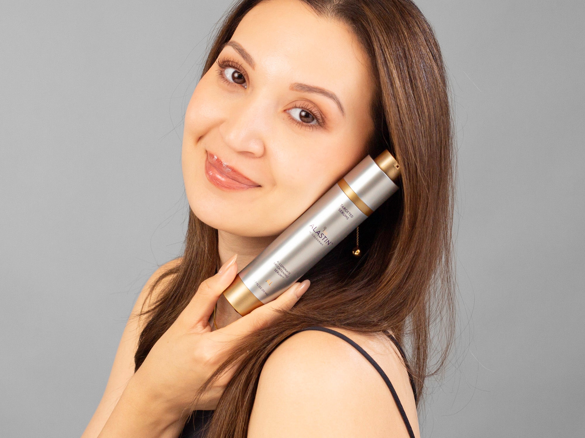 Laziza from LAZ Skincare holding a bottle of Alastin A-Luminate Brightening Serum against a gray background