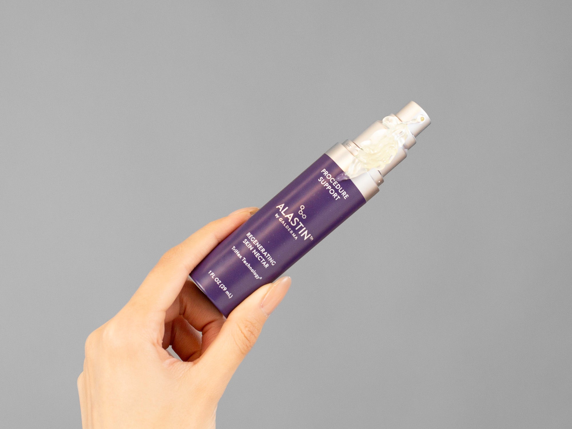 Laziza from LAZ Skincare's hand holding a purple Alastin Regenerating Skin Nectar bottle against a gray background